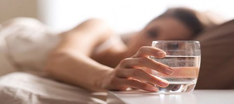 Eine junge Dame liegt im Bett und greift nach einem Glas Wasser auf dem Nachttisch, weil sie Nachts weniger schwitzen will.